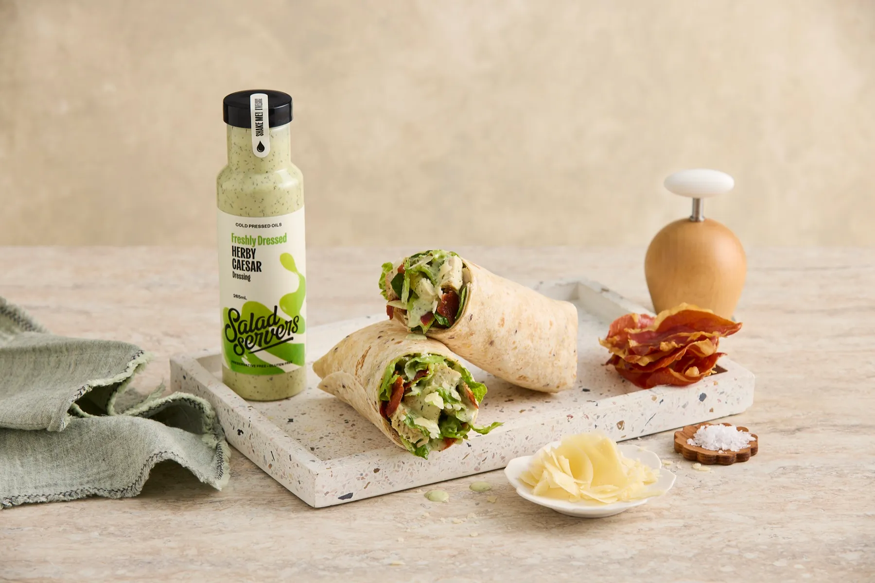 Chicken Caesar wrap cut in half on a terrazzo board, beside a bottle of Salad Servers Herby Caesar dressing, crispy prosciutto and shaved parmesan