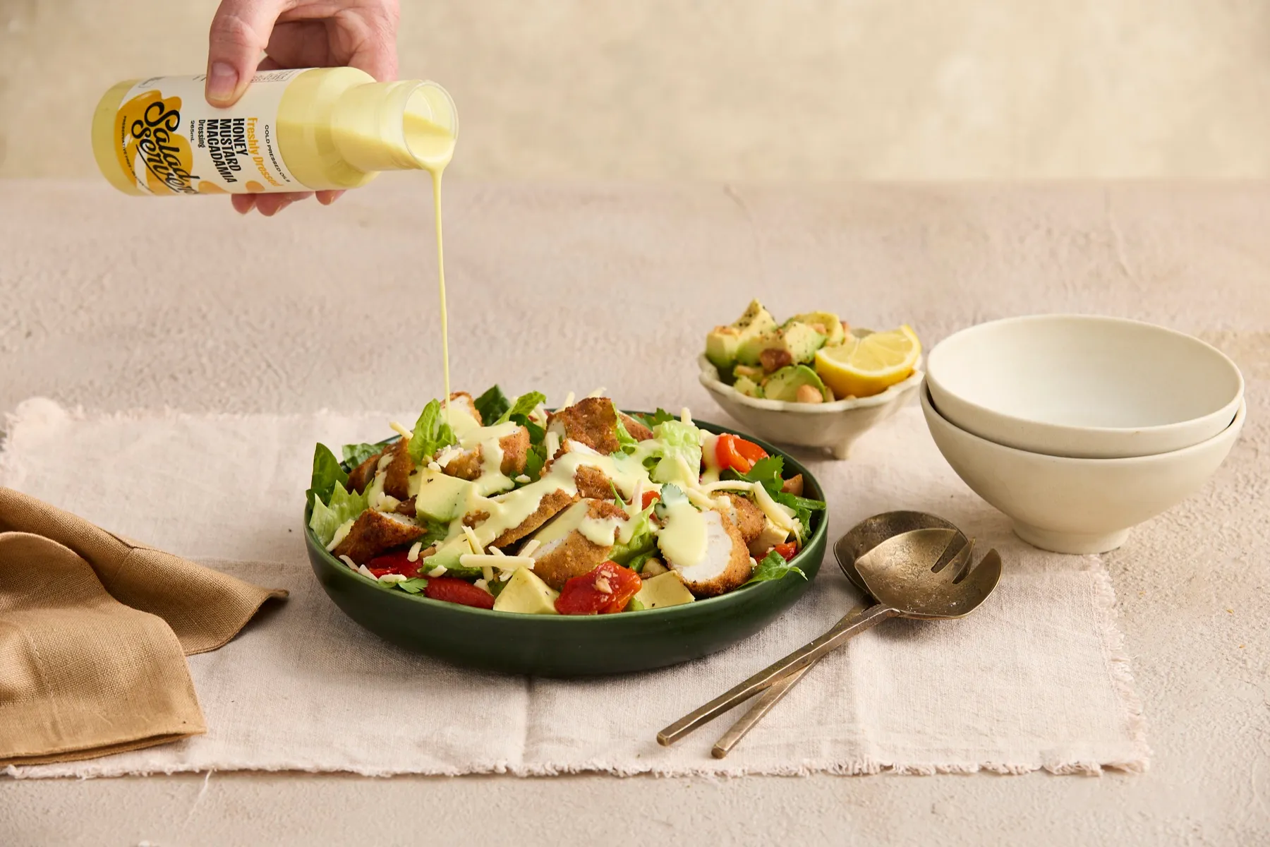 Salad Servers Honey Mustard Macadamia dressing being poured over a bowl of crumbed chicken, cos lettuce, avocado and roast capsicum